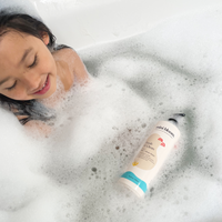 A smiling girl relaxes in a bubble bath with Mini Bloom Fresh N' Clean hair and body wash.

