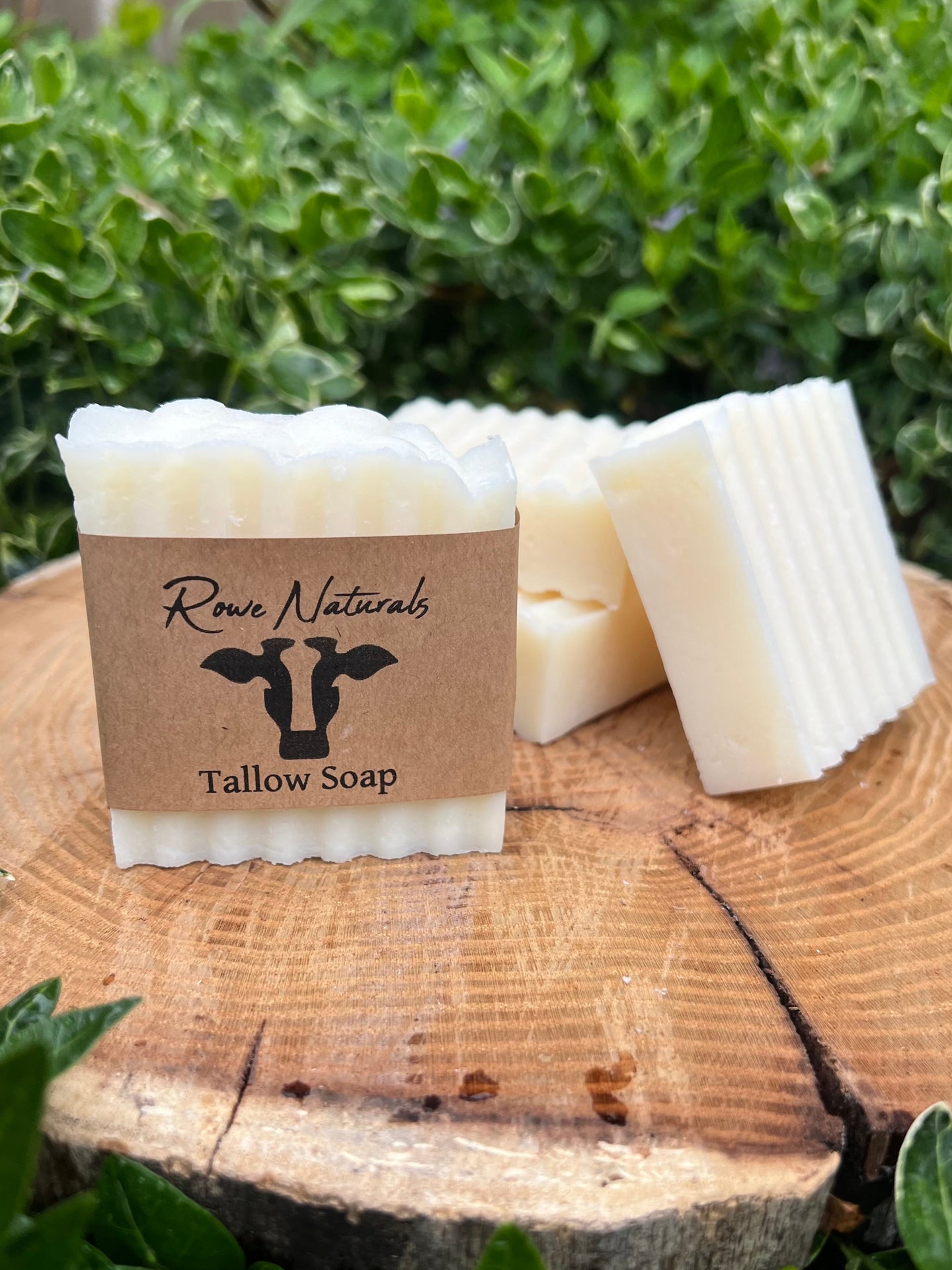 Three bars of Rowe Naturals tallow soap rest on a wood slice against a green leafy background.
