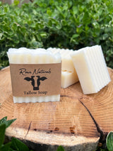 Three bars of Rowe Naturals tallow soap rest on a wood slice against a green leafy background.
