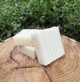 Three bars of textured, handmade soap rest on a rustic wooden surface against a leafy green backdrop.
