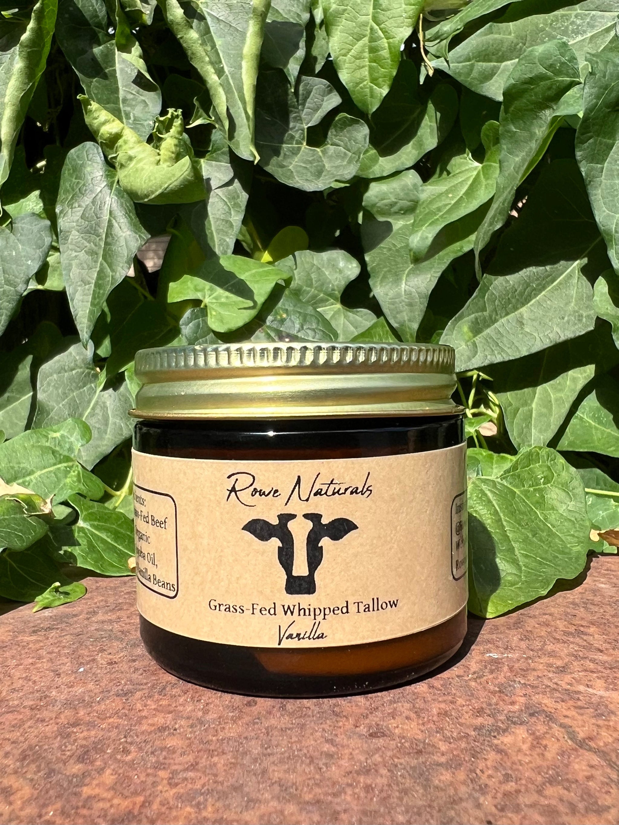 Rowe Naturals sells grass-fed whipped tallow vanilla balm in a glass jar with lush green foliage behind it.
