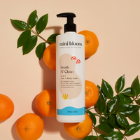 Mini Bloom's Fresh N' Clean 2-in-1 hair and body wash sits among oranges and foliage, highlighting its natural ingredients.
