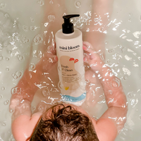 A baby holds a bottle of mini bloom Fresh N' Clean 2-in-1 hair and body wash while sitting in a bubble bath.

