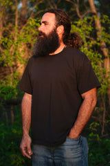 Bearded man with long hair and black t-shirt gazes thoughtfully into the distance outdoors.
