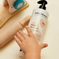 A child's hand reaches for a bottle of Mini Bloom Knots Be Gone hair detangler, displayed with a wooden rolling pin and toy scale.
