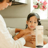 A parent bathes a toddler with Mini Bloom "Knots Be Gone" hair detangling conditioner in a bright bathroom.
