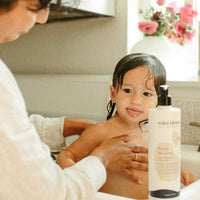 A parent bathes a toddler with Mini Bloom "Knots Be Gone" hair detangling conditioner in a bright bathroom.
