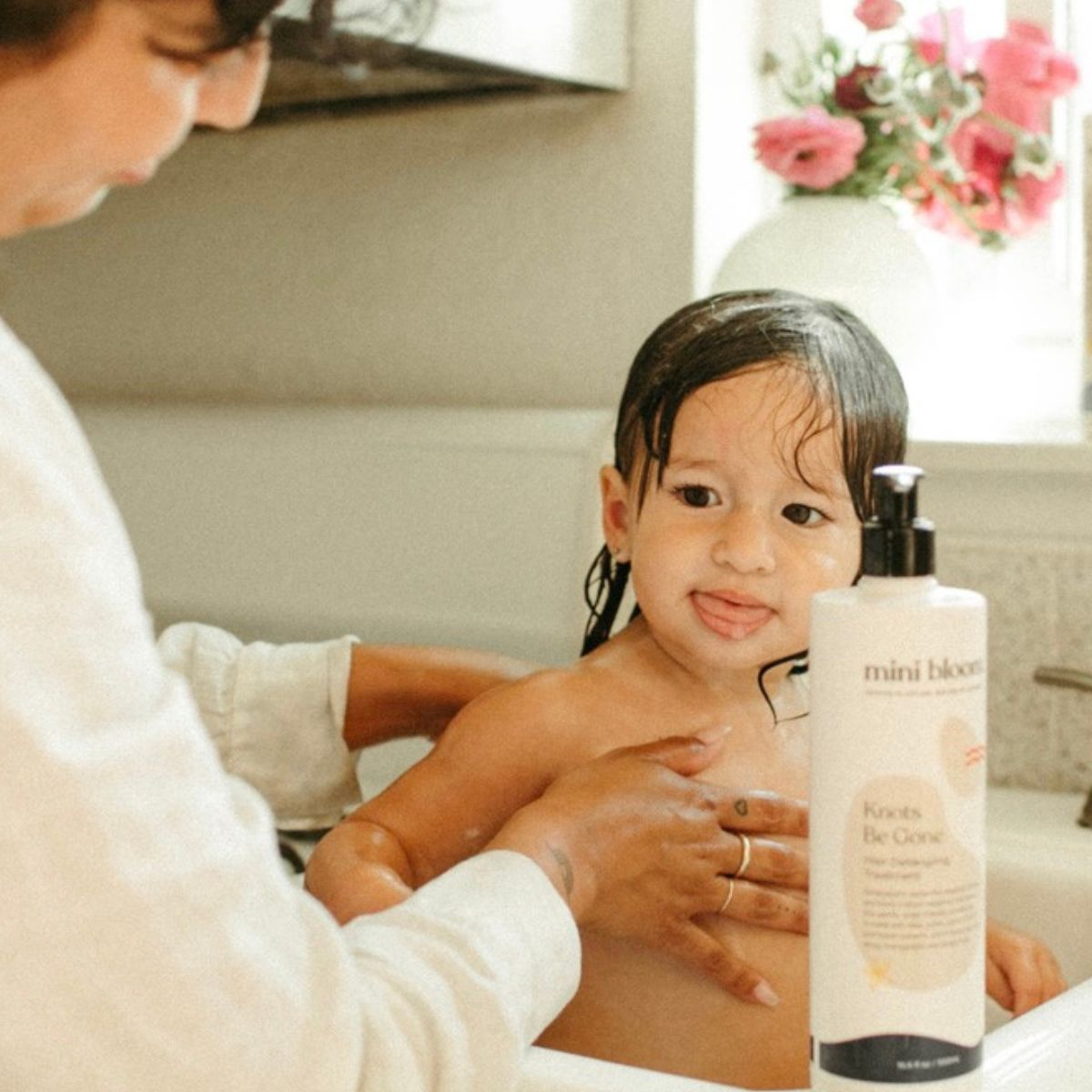 A parent bathes a toddler with Mini Bloom "Knots Be Gone" hair detangling conditioner in a bright bathroom.

