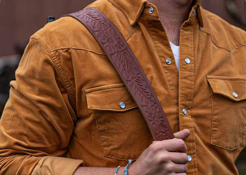 A close-up of a person wearing a brown leather crossbody strap with an embossed floral pattern over a mustard brown corduroy button-up shirt.