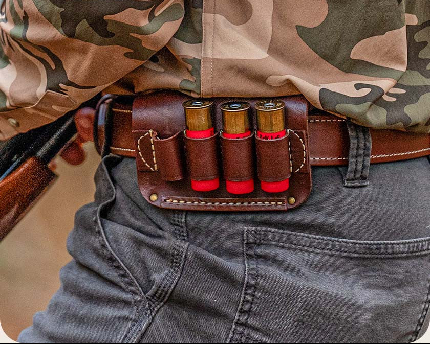A close-up of a brown leather shotgun shell pouch attached to a brown belt, worn by a person in a camouflage shirt and gray jeans.