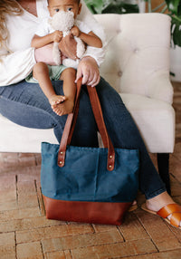 Woman holding a blue and brown leather tote bag sits with her child hugging a stuffed animal.
