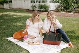 Two friends enjoy a stylish summer picnic with flowers, cheese, and bread on a blanket in the grass.
