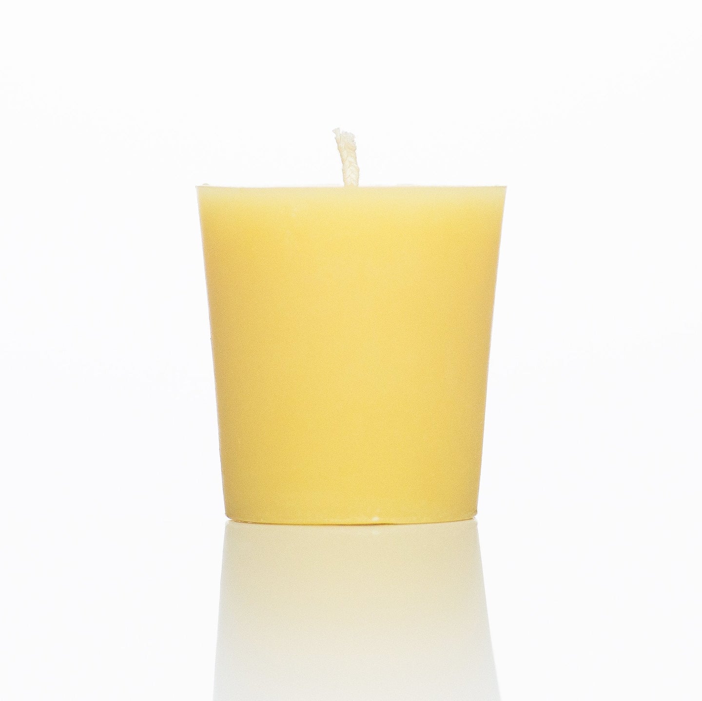 A natural beeswax votive candle sits on a reflective white surface.
