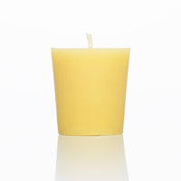 A natural beeswax votive candle sits on a reflective white surface.
