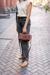 A woman showcases her stylish fall outfit, featuring a brown leather satchel bag, grey jeans, a cream sweater, and tan loafers, on a brick sidewalk.
