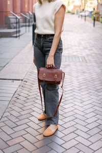A woman showcases her stylish fall outfit, featuring a brown leather satchel bag, grey jeans, a cream sweater, and tan loafers, on a brick sidewalk.
