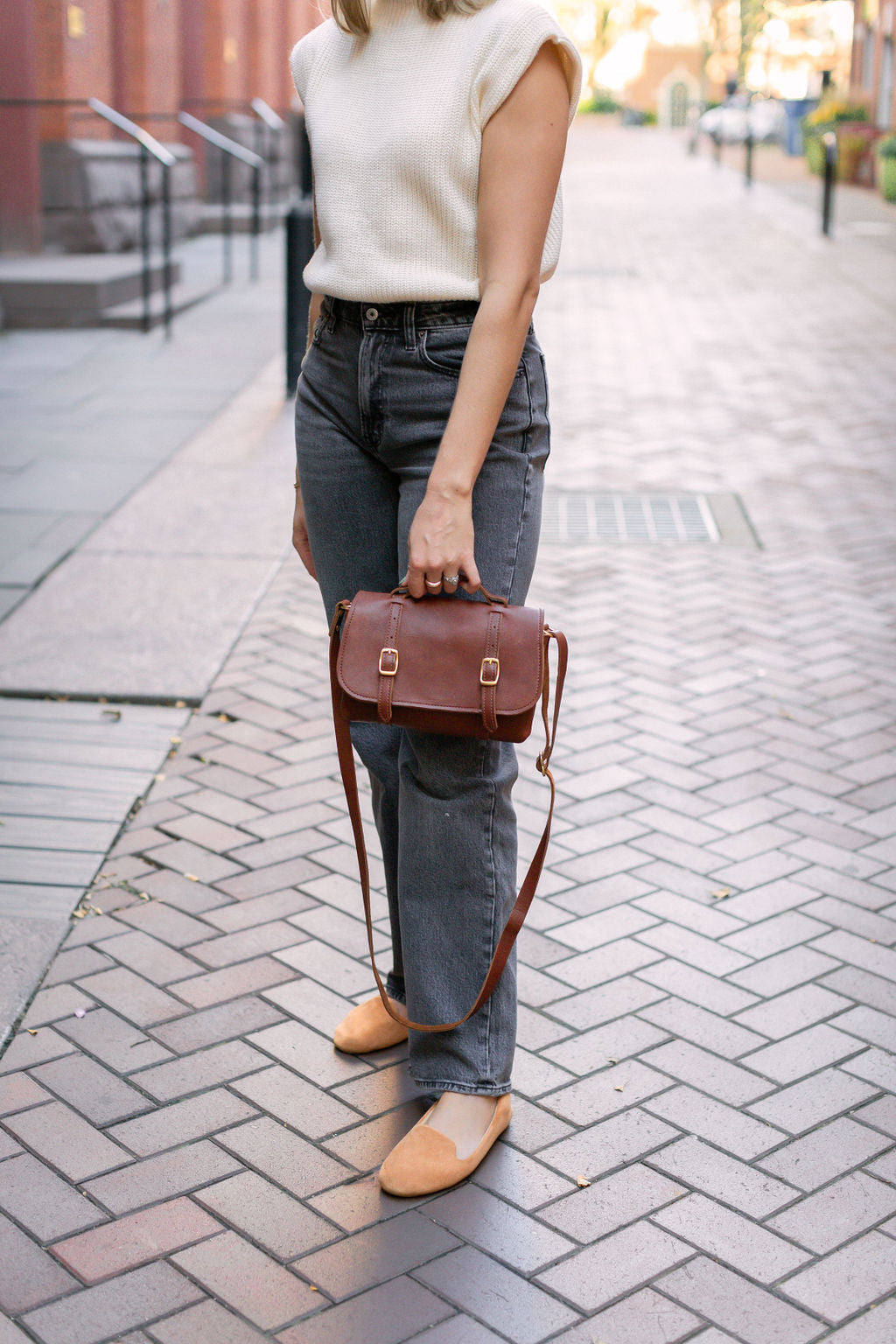 A woman showcases her stylish fall outfit, featuring a brown leather satchel bag, grey jeans, a cream sweater, and tan loafers, on a brick sidewalk.
