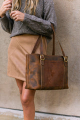 A woman models a brown leather tote bag with gold hardware, worn with a gray sweater and tan skirt.
