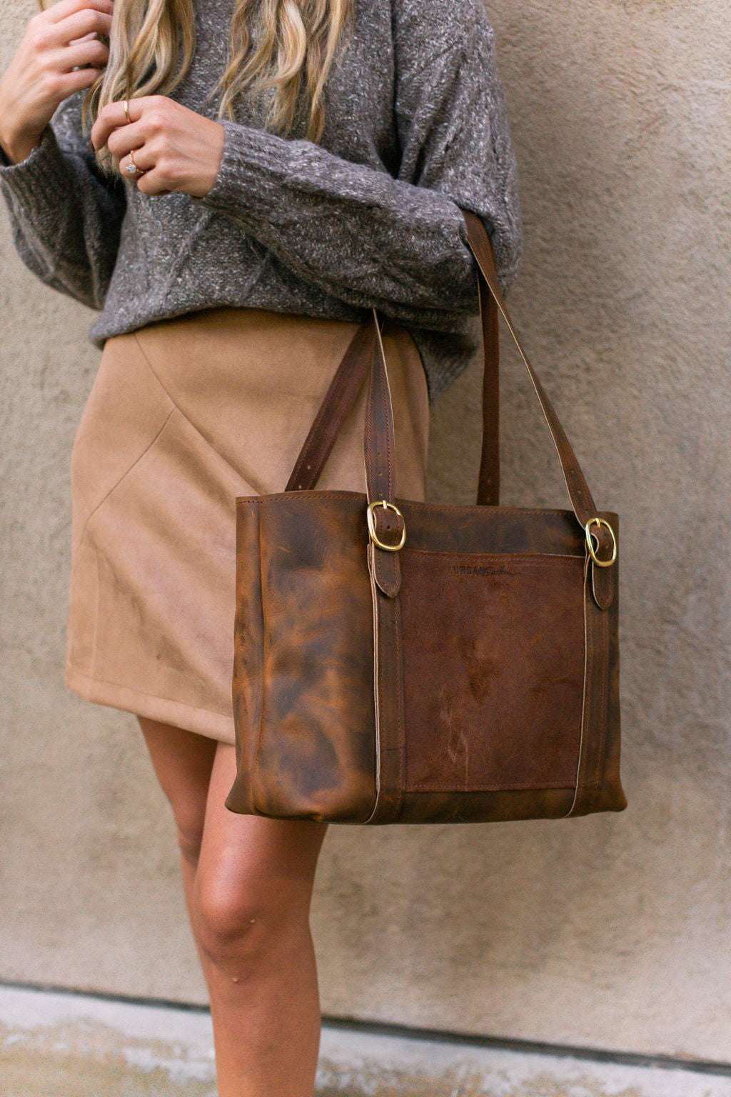 A woman models a brown leather tote bag with gold hardware, worn with a gray sweater and tan skirt.
