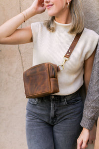 A blonde woman models a brown leather crossbody bag with a gold clasp and strap while wearing a cream sweater vest and gray jeans.
