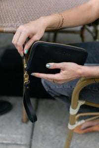 Woman unzips a black leather wristlet wallet with gold hardware while sitting outdoors.
