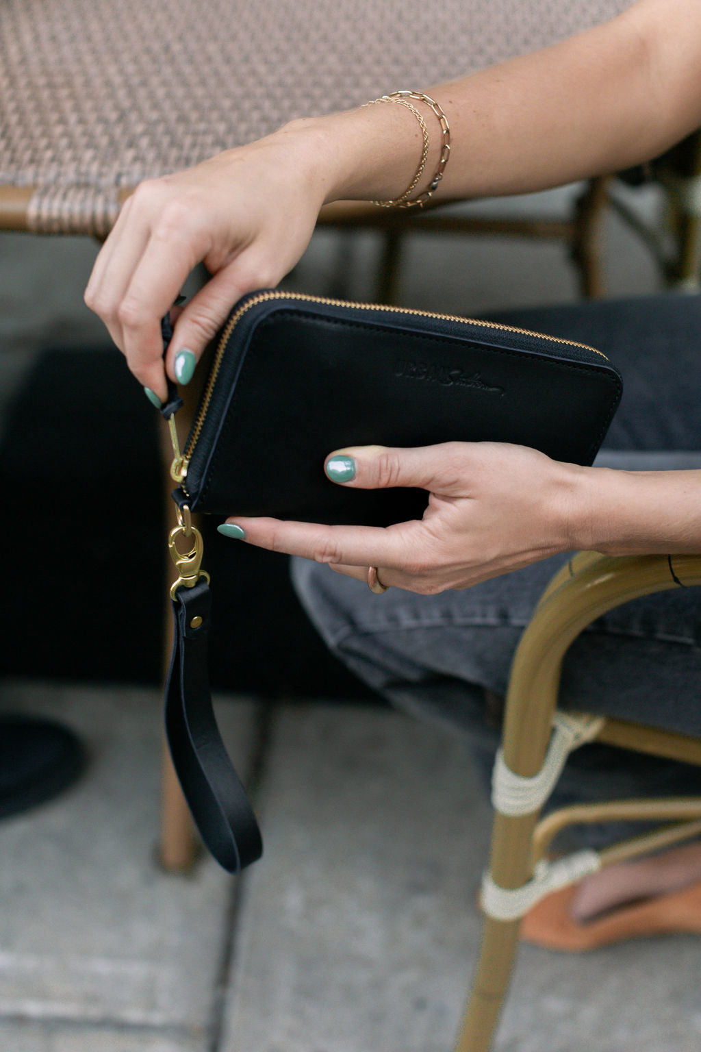 Woman unzips a black leather wristlet wallet with gold hardware while sitting outdoors.
