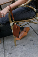 Woman sits on a rattan chair with a brown leather wallet bag hanging from the side, wearing gray jeans and tan suede flats.
