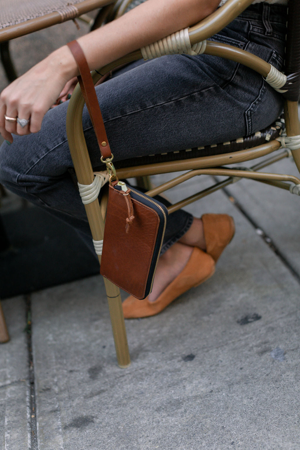 Woman sits on a rattan chair with a brown leather wallet bag hanging from the side, wearing gray jeans and tan suede flats.
