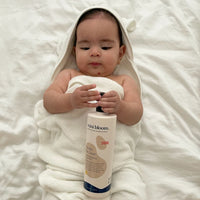 A baby wearing a hooded towel examines a bottle of Mini Bloom "Knots Be Gone" detangling treatment.
