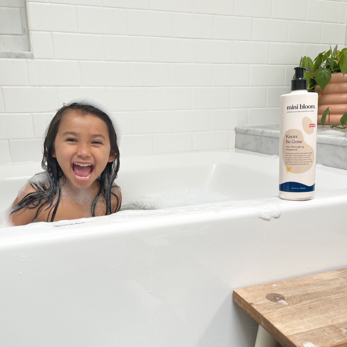 A happy young girl smiles in a bubble-filled bathtub, next to a bottle of Mini Bloom "Knots Be Gone" hair detangler.
