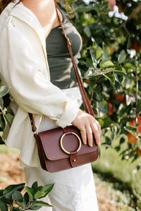 Woman models a stylish brown leather crossbody bag with gold hardware while standing by an apple tree.
