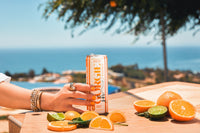 A hand holding a Jaclyn Hill's Orange Twist Gorgie Energy drink poses on a table decorated with oranges and limes with an ocean view.
