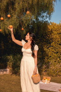A woman in a white dress picks fresh oranges from a tree during a summer picnic in a sun-drenched orchard.