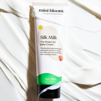Mini Bloom Silk Milk Baby Cream sits on a background of creamy white lotion.
