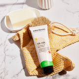 Mini Bloom Silk Milk baby cream sits beside a bar of soap, a brush, and a yellow washcloth, showcasing skincare for infants.
