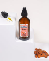 VanMan's Protection Oils bottle sits on a white step next to almonds, with a dropper showing the oil's golden color.

