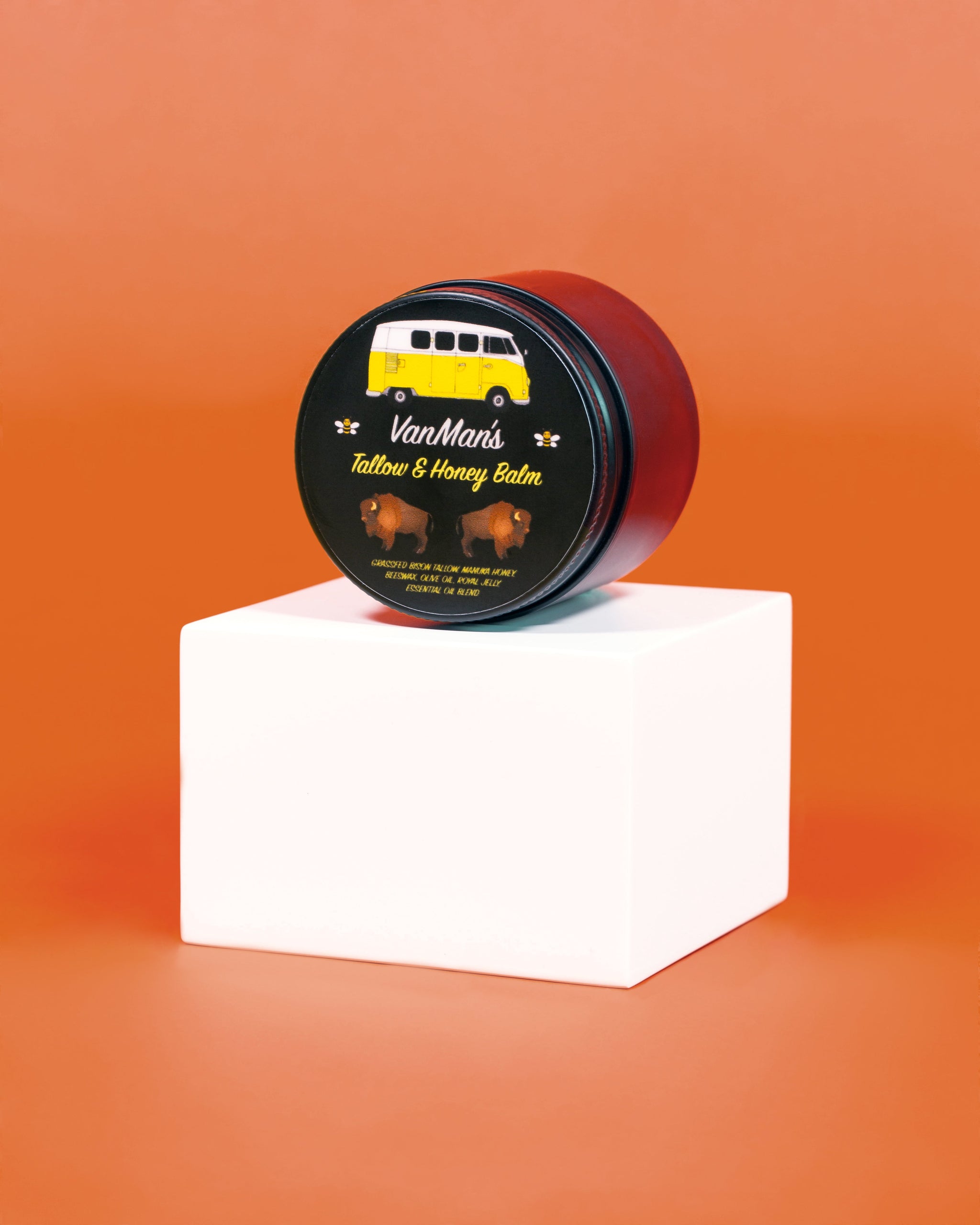 VanMan's Tallow & Honey Balm tin, featuring a vintage van and bison illustrations, sits on a white cube against an orange background, showcasing natural skincare ingredients like grass-fed bison tallow and Manuka honey.
