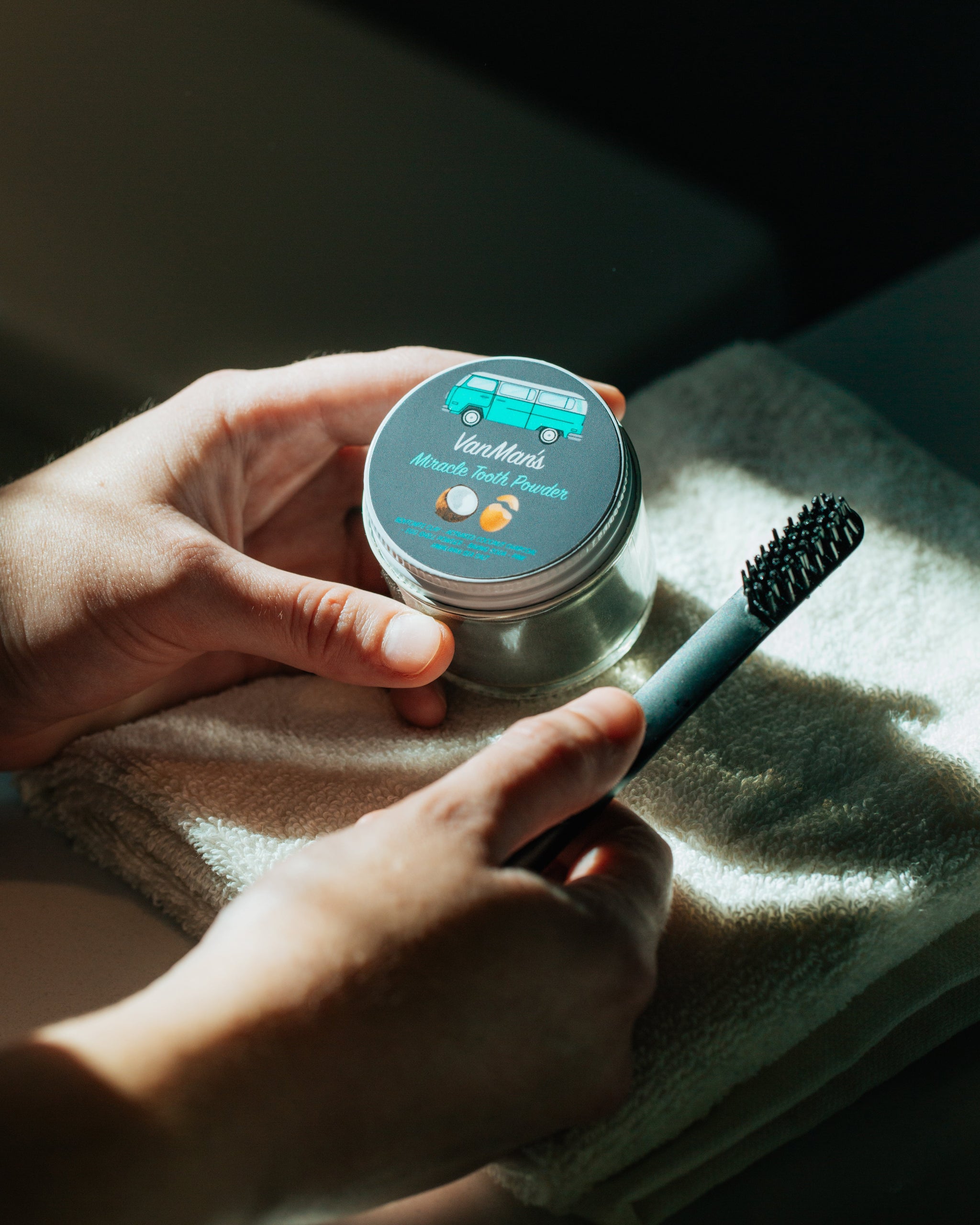 Hands hold VanMan's Miracle Tooth Powder and a bamboo toothbrush over a white towel in natural light.
