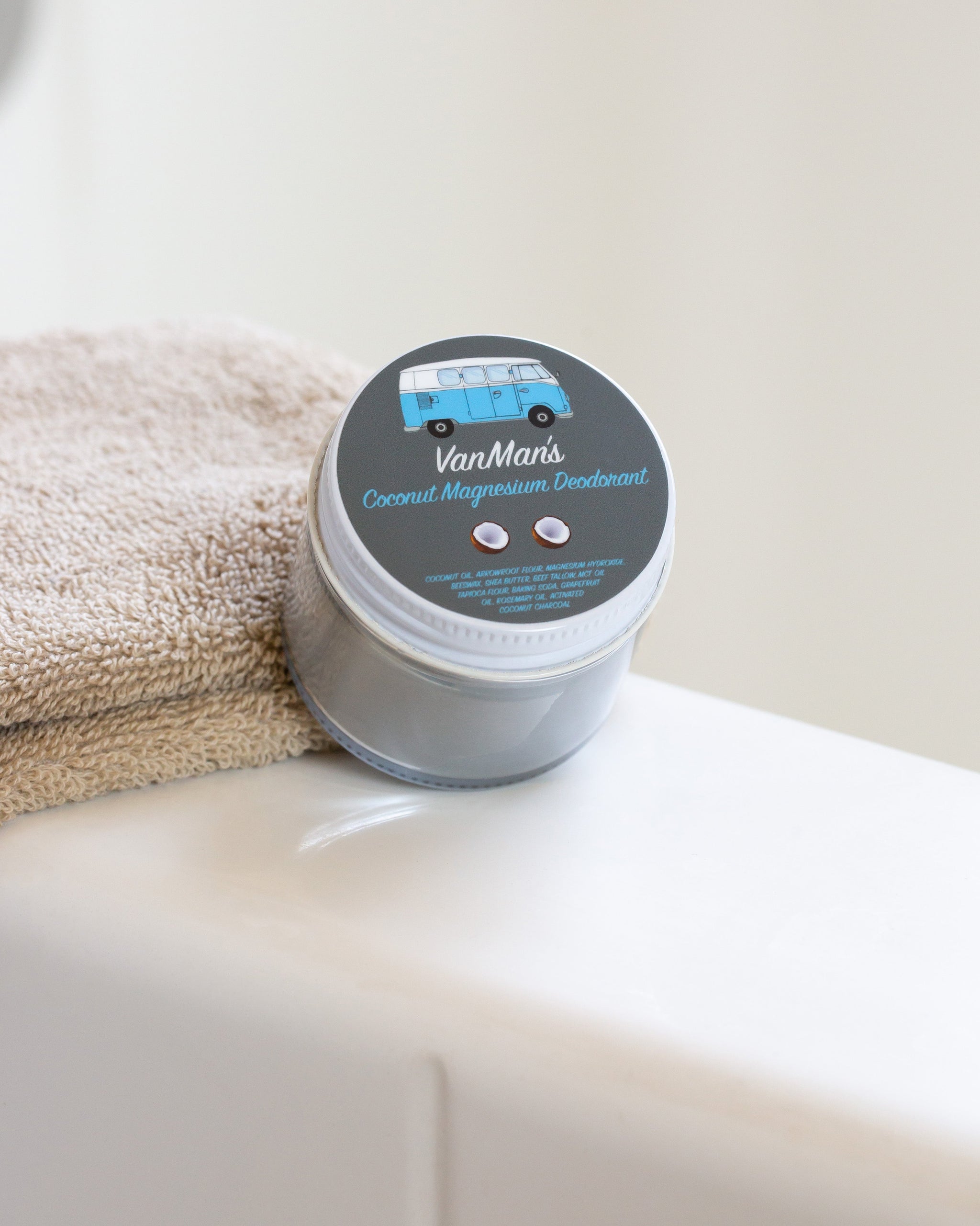 VanMan's coconut magnesium deodorant, featuring a VW bus illustration, sits on a bathroom sink next to a folded towel.
