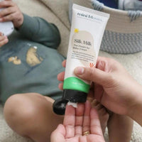 Someone dispenses Mini Bloom Silk Milk baby cream onto their hand near a child.
