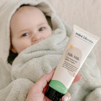 Hand displays Mini Bloom Silk Milk Baby Cream, with a baby wrapped in a towel visible in the background.
