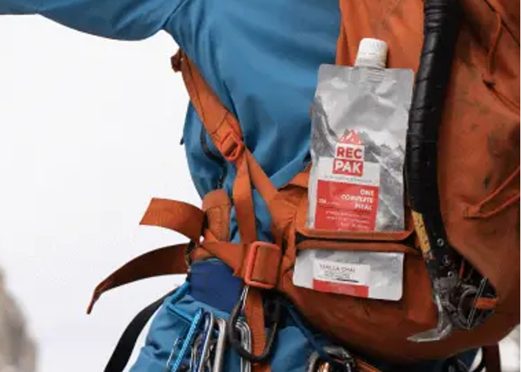 A close-up of a hiker wearing a blue jacket and black pants, carrying an orange backpack with climbing gear and a RecPak attached to the side harness, against a snowy mountain background.