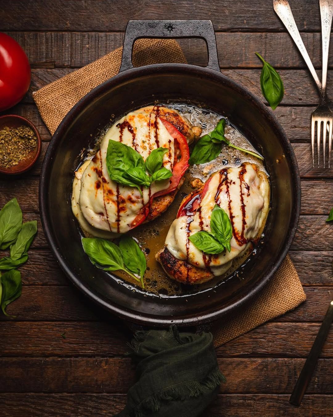 Grilled chicken breast topped with melted mozzarella cheese, balsamic glaze, and fresh basil leaves, served in a dark cast-iron skillet on a rustic wooden table with a tomato, herbs, and cutlery around it.