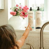A child reaches for Mini Bloom kids' Fresh N' Clean body wash and Knots Be Gone detangler displayed on a window sill with pink flowers.
