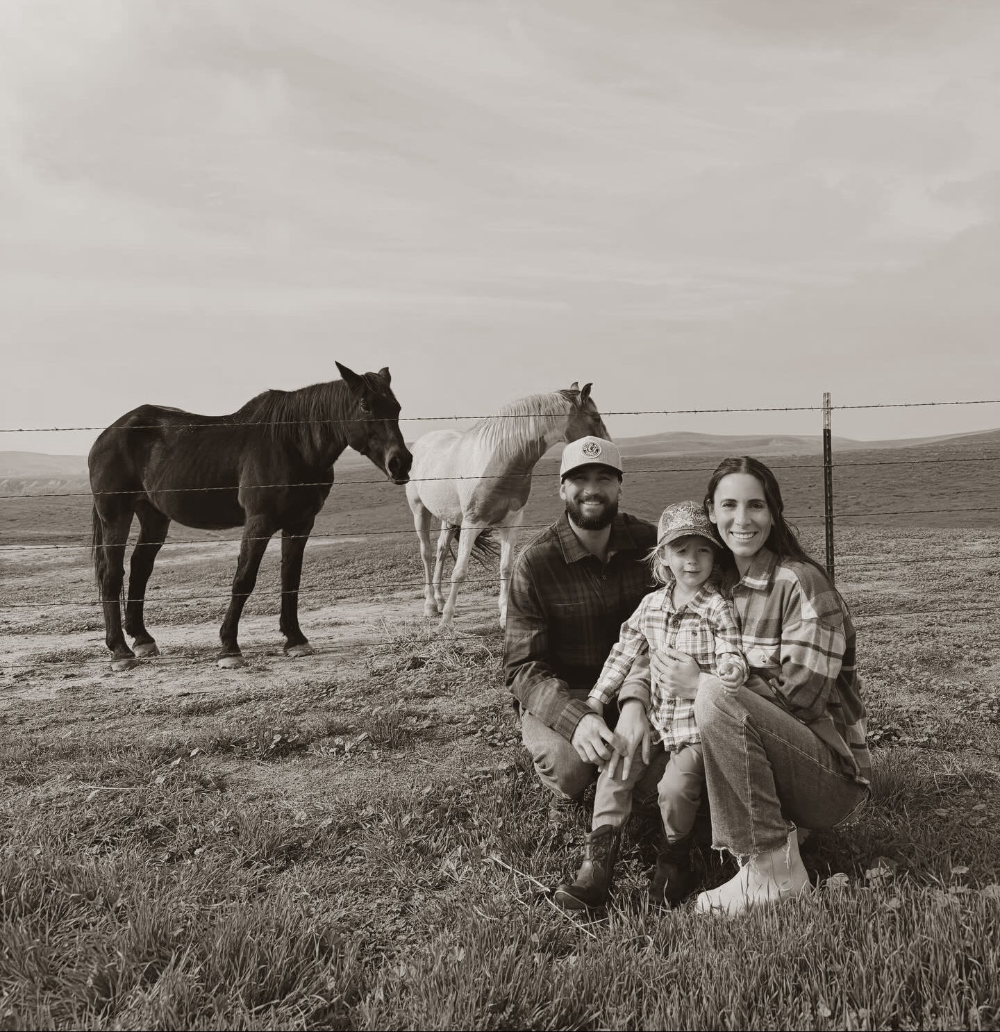 A sepia-toned family portrait showing a man, a woman, and a young child kneeling in a grassy field with two horses behind them. The photo has a warm monochrome, vintage aesthetic.