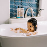A happy little girl plays with bubbles in her bathtub next to Mini Bloom kids' bath products.
