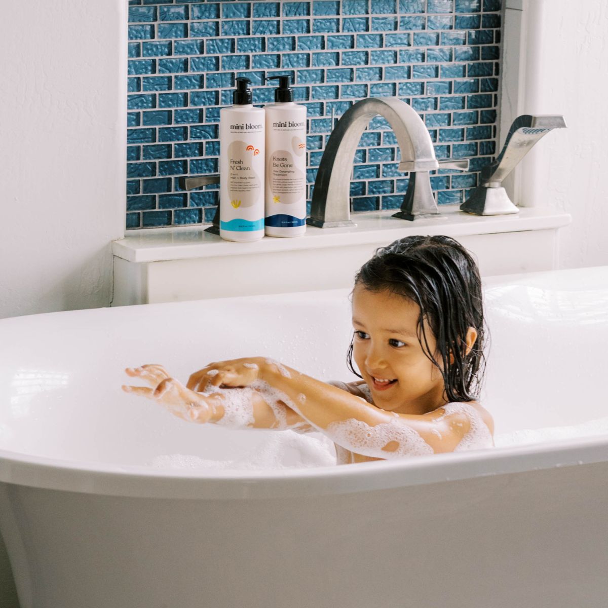A happy little girl plays with bubbles in her bathtub next to Mini Bloom kids' bath products.
