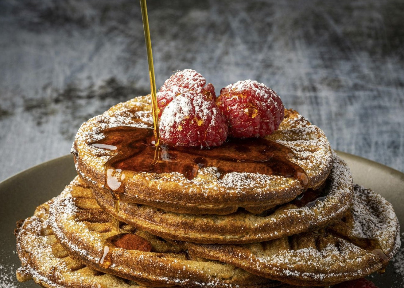 A stack of golden-brown pancakes topped with fresh raspberries, powdered sugar, and a drizzle of syrup, served on a gray plate with a fork on a dark rustic table.