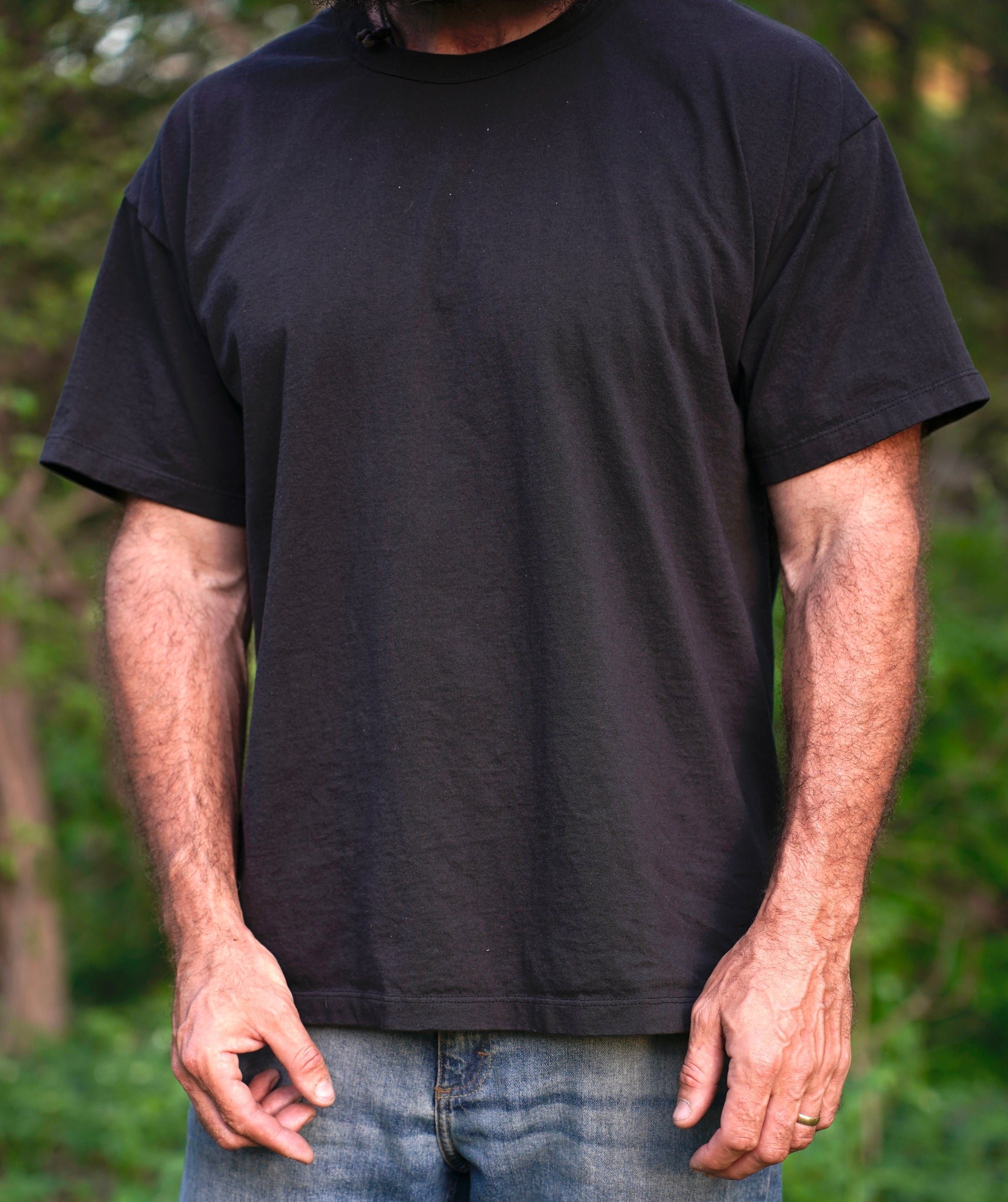 Man models a blank black t-shirt and denim jeans against a blurred green outdoor background.
