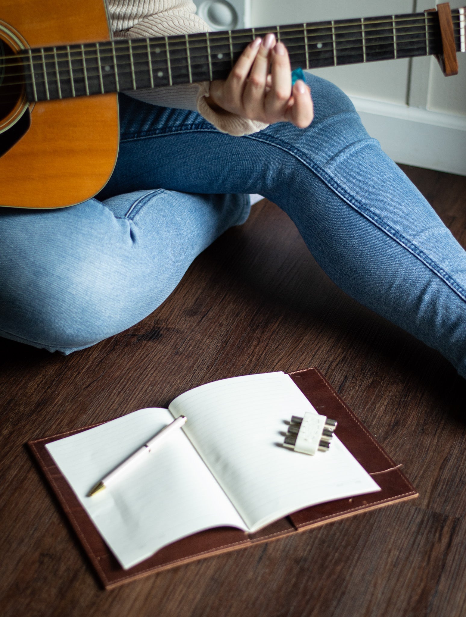 A musician in jeans strums an acoustic guitar while sitting on the floor next to an open notebook and pen, ready for songwriting.

